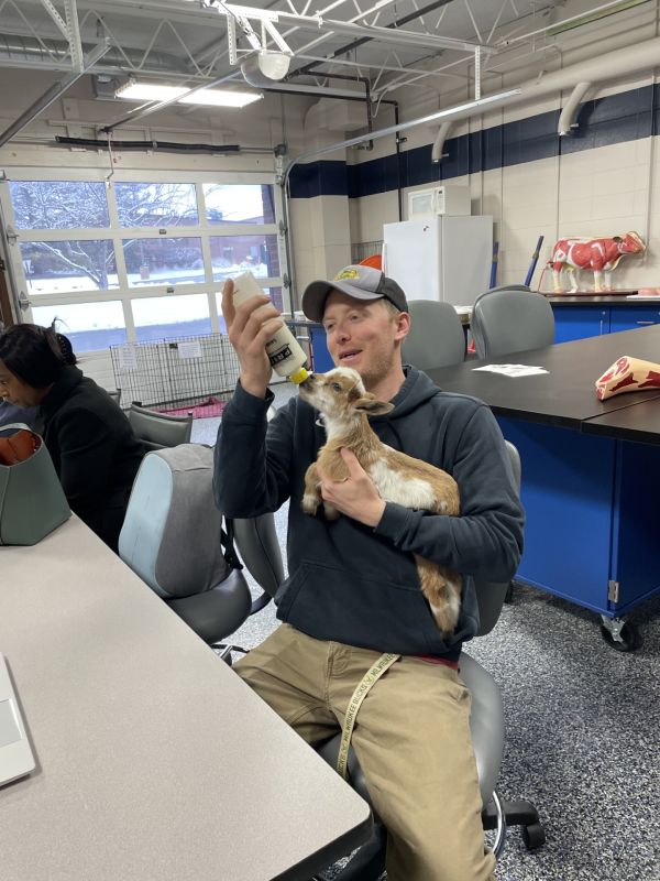 Participant in black hoodie and a baseball cap is holding a baby goat and feeding it from a bottle inside a room with large garage doors.